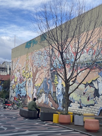 A vibrant graffiti mural covers a large wall, featuring a mix of abstract shapes, figures, and symbols in various bright colors. In front of the mural, a leafless tree is prominent, and a person in a green jacket and black hat sits on a concrete bench. Several colorful planters and a few bicycles are also visible on the patterned pavement.