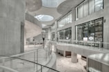 The image depicts a modern, spacious interior with curved architectural features and large glass windows. The space is characterized by sleek, white surfaces and minimalist design elements. There are a few people visible in the distance, adding a sense of scale to the large, open area.