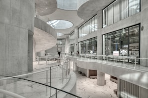 The image depicts a modern, spacious interior with curved architectural features and large glass windows. The space is characterized by sleek, white surfaces and minimalist design elements. There are a few people visible in the distance, adding a sense of scale to the large, open area.