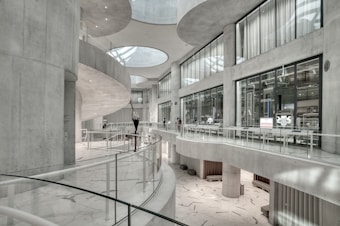 The image depicts a modern, spacious interior with curved architectural features and large glass windows. The space is characterized by sleek, white surfaces and minimalist design elements. There are a few people visible in the distance, adding a sense of scale to the large, open area.