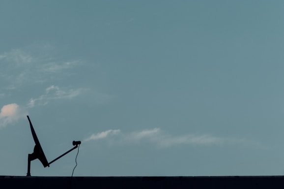 A silhouette of a satellite dish with a cable is positioned on a flat surface against a clear blue sky. A few faint clouds are visible, creating a serene and minimalistic scene.