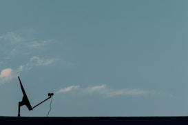 A silhouette of a satellite dish with a cable is positioned on a flat surface against a clear blue sky. A few faint clouds are visible, creating a serene and minimalistic scene.