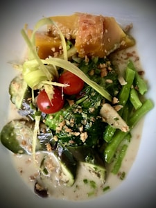 A vibrant dish featuring a combination of colorful vegetables such as cherry tomatoes, sliced eggplant, and green beans. The dish is garnished with crispy, golden bits and drizzled with a creamy, light sauce. The texture is enhanced with thin strips of a pale green garnish laid over the top.