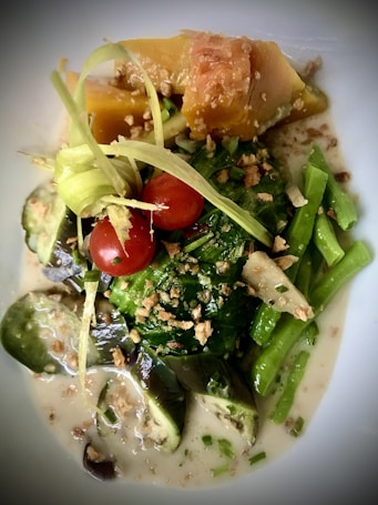 A vibrant dish featuring a combination of colorful vegetables such as cherry tomatoes, sliced eggplant, and green beans. The dish is garnished with crispy, golden bits and drizzled with a creamy, light sauce. The texture is enhanced with thin strips of a pale green garnish laid over the top.