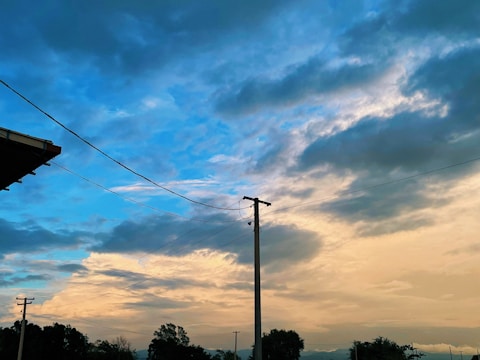 A dynamic sky blends hues of blue and orange, with clouds adding texture and depth. Power lines intersect the image, leading to a utility pole, while trees form a dark silhouette against the bright sky.