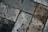 A close-up view of textured paving stones arranged in a diagonal pattern. The stones display varying shades of gray and brown, with visible wear, stains, and slight weathering, enhancing the rough texture and rustic appearance.