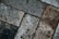 A close-up view of textured paving stones arranged in a diagonal pattern. The stones display varying shades of gray and brown, with visible wear, stains, and slight weathering, enhancing the rough texture and rustic appearance.