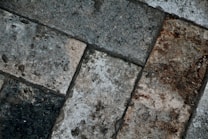 A close-up view of textured paving stones arranged in a diagonal pattern. The stones display varying shades of gray and brown, with visible wear, stains, and slight weathering, enhancing the rough texture and rustic appearance.