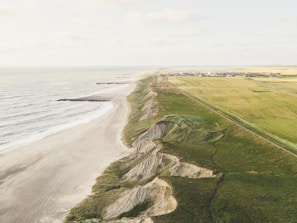 An aerial view of a coastline featuring a sandy beach on the left, rugged cliffs in the middle, and expansive agricultural fields on the right. In the distance, there is a small settlement with houses and buildings.