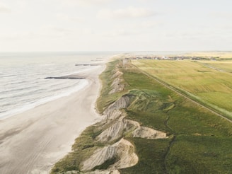 An aerial view of a coastline featuring a sandy beach on the left, rugged cliffs in the middle, and expansive agricultural fields on the right. In the distance, there is a small settlement with houses and buildings.