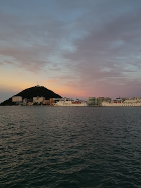A calm harbor scene with cargo ships docked under a clear sky at sunset.