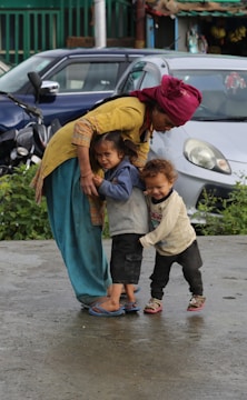 A woman wearing a red headscarf and yellow top is leaning over towards two young children, one of whom is holding onto her leg. They are on a wet concrete surface with parked vehicles and greenery in the background.