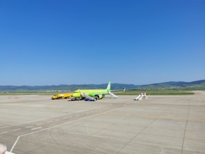 An airport apron featuring a lime-green airplane parked on the tarmac. Nearby, a yellow service vehicle and aircraft stairs are positioned. The background reveals grassy hills under a clear blue sky, with mountains in the distance.