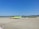 An airport apron featuring a lime-green airplane parked on the tarmac. Nearby, a yellow service vehicle and aircraft stairs are positioned. The background reveals grassy hills under a clear blue sky, with mountains in the distance.
