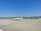An airport apron featuring a lime-green airplane parked on the tarmac. Nearby, a yellow service vehicle and aircraft stairs are positioned. The background reveals grassy hills under a clear blue sky, with mountains in the distance.