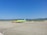 An airport apron featuring a lime-green airplane parked on the tarmac. Nearby, a yellow service vehicle and aircraft stairs are positioned. The background reveals grassy hills under a clear blue sky, with mountains in the distance.