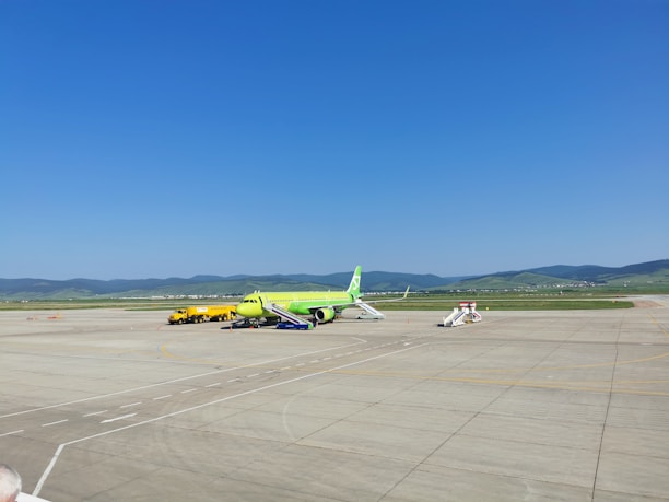 An airport apron featuring a lime-green airplane parked on the tarmac. Nearby, a yellow service vehicle and aircraft stairs are positioned. The background reveals grassy hills under a clear blue sky, with mountains in the distance.