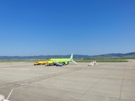 An airport apron featuring a lime-green airplane parked on the tarmac. Nearby, a yellow service vehicle and aircraft stairs are positioned. The background reveals grassy hills under a clear blue sky, with mountains in the distance.