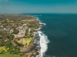 A coastal landscape with a vast expanse of blue ocean meeting the shoreline, lined with waves crashing against the rocks. A small town is visible with numerous buildings and a mix of green and brown land areas, indicating vegetation and urban development.