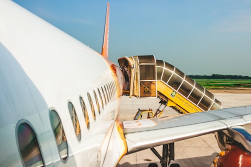 An airplane is parked on the runway with a boarding ramp connected to one of its doors. The aircraft is white with orange accents, and the ramp is labeled 'SAIGON GROUND SERVICE'. The scene is set against a clear blue sky and a vast expanse of pavement.