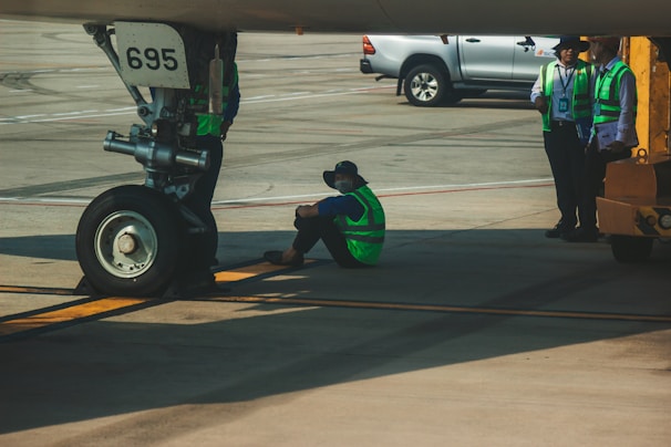 An aircraft is parked on the tarmac with a visible nose landing gear marked with the number 695. A person wearing a green safety vest and a cap is sitting on the ground near the landing gear, possibly taking a break. Three other individuals, also in safety vests, are standing nearby along with a vehicle parked in the background. The setting appears to be an airport runway or hangar area.