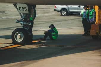 An aircraft is parked on the tarmac with a visible nose landing gear marked with the number 695. A person wearing a green safety vest and a cap is sitting on the ground near the landing gear, possibly taking a break. Three other individuals, also in safety vests, are standing nearby along with a vehicle parked in the background. The setting appears to be an airport runway or hangar area.
