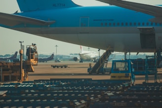 The image depicts a busy airport apron with multiple airplanes visible. One large aircraft with the identifier 'HL7714' is prominent in the foreground. Various ground support equipment including a yellow vehicle and baggage carts are also visible, with multiple aircraft in the background preparing for departure or arrival.