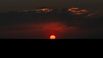 The image features a sunset with a deep red sun half-submerged below the horizon, surrounded by dark clouds with hints of orange highlighting the cloud edges. The sky transitions from a vivid red near the sun to a darker blue-black as you move upwards.