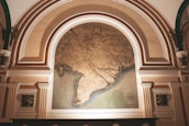 An ornate archway features a large, detailed map with intricate lines and coastal outlines. The map is framed by architectural elements with decorative columns and embellishments, suggesting a historic or museum setting. The map itself appears aged, with a color palette of earthy tones.