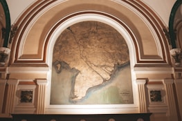An ornate archway features a large, detailed map with intricate lines and coastal outlines. The map is framed by architectural elements with decorative columns and embellishments, suggesting a historic or museum setting. The map itself appears aged, with a color palette of earthy tones.