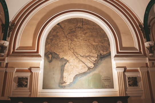 An ornate archway features a large, detailed map with intricate lines and coastal outlines. The map is framed by architectural elements with decorative columns and embellishments, suggesting a historic or museum setting. The map itself appears aged, with a color palette of earthy tones.