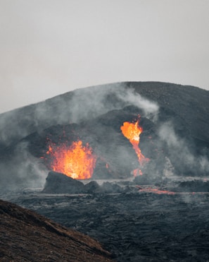A volcanic eruption with molten lava flowing from the crater, surrounded by smoke and ash. The lava is bright orange and red against the dark, charred landscape. Plumes of white smoke rise into the grey sky, creating a dramatic and intense scene.