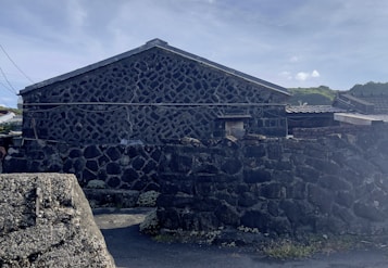 The image depicts a traditional stone house with a gabled roof. The walls and perimeter of the property are constructed from rough, dark stones, giving it a rustic appearance. In the background, there are low-lying hills and a partly cloudy sky. Electrical wires run across the fa&ccedil;ade of the building.