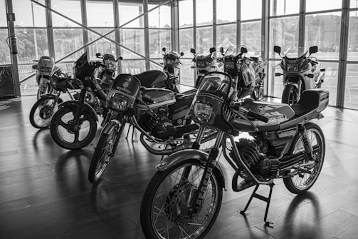 A collection of vintage mopeds is displayed in a showroom with large windows, allowing natural light to illuminate the space. The bikes are positioned in neat rows on a wooden floor, creating a sense of nostalgia and admiration for classic motorbikes.