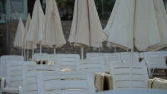 A collection of closed beige patio umbrellas standing amid numerous white plastic lounge chairs, set against a stone wall backdrop in an outdoor environment.