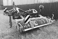 A piece of agricultural machinery with a metal frame and cylindrical attachment is situated on a gravel and grassy surface. The equipment appears to be used for soil preparation, possibly a cultivator or similar farming tool. It is positioned near a corrugated metal building or fence.