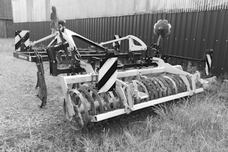 A piece of agricultural machinery with a metal frame and cylindrical attachment is situated on a gravel and grassy surface. The equipment appears to be used for soil preparation, possibly a cultivator or similar farming tool. It is positioned near a corrugated metal building or fence.