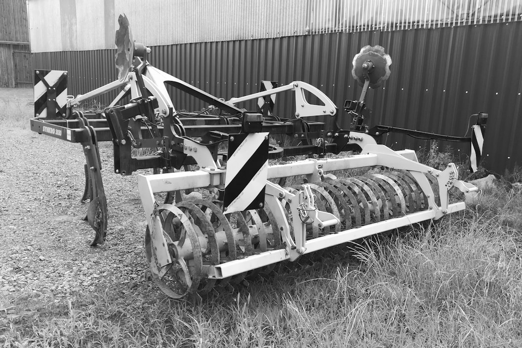 A piece of agricultural machinery with a metal frame and cylindrical attachment is situated on a gravel and grassy surface. The equipment appears to be used for soil preparation, possibly a cultivator or similar farming tool. It is positioned near a corrugated metal building or fence.