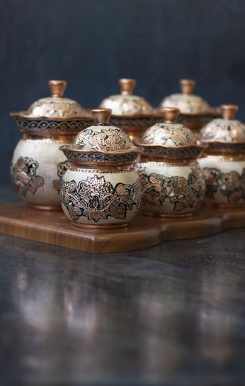 A collection of ornately decorated ceramic pots with lids, featuring intricate floral patterns. The pots are placed on a wooden tray, with a dark, blurred background that emphasizes the craftsmanship and detail of the pottery.