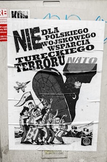 A poster with bold text in Polish related to political or military issues, featuring a black and white illustration of a large animal, possibly a bull, with human figures and a vehicle or military aircraft. The figures are depicted with expressive gestures, suggesting a theme of protest or opposition.