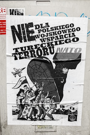A poster with bold text in Polish related to political or military issues, featuring a black and white illustration of a large animal, possibly a bull, with human figures and a vehicle or military aircraft. The figures are depicted with expressive gestures, suggesting a theme of protest or opposition.