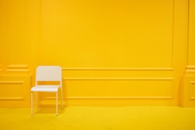 A minimalist scene featuring a single white chair placed against a vibrant yellow wall with paneling details. The floor is also yellow, creating a monochromatic and bold aesthetic.