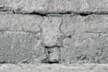 A close-up view of a textured concrete wall featuring horizontal lines and rough, granular details. The surface appears weathered and uneven, with visible cracks and variations in the stone.