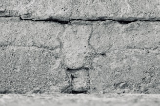 A close-up view of a textured concrete wall featuring horizontal lines and rough, granular details. The surface appears weathered and uneven, with visible cracks and variations in the stone.