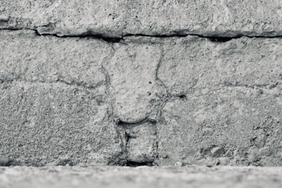 A close-up view of a textured concrete wall featuring horizontal lines and rough, granular details. The surface appears weathered and uneven, with visible cracks and variations in the stone.