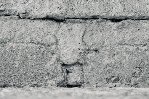 A close-up view of a textured concrete wall featuring horizontal lines and rough, granular details. The surface appears weathered and uneven, with visible cracks and variations in the stone.