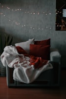 A cozy armchair draped with soft blankets and cushions, surrounded by a string of warm fairy lights. The setting is likely a living room with a dark, textured wall and a potted plant nearby.
