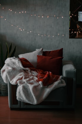 A cozy armchair draped with soft blankets and cushions, surrounded by a string of warm fairy lights. The setting is likely a living room with a dark, textured wall and a potted plant nearby.