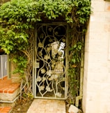 A wrought iron gate decorated with leaf patterns is set within a lush wall of green foliage. The gate leads to a pathway made of stone, which is partially visible through the opening. The surrounding structure includes a wall of light-colored stone and red brick steps.