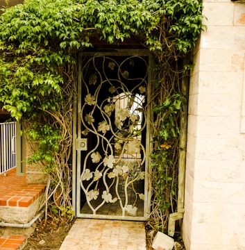 A wrought iron gate decorated with leaf patterns is set within a lush wall of green foliage. The gate leads to a pathway made of stone, which is partially visible through the opening. The surrounding structure includes a wall of light-colored stone and red brick steps.